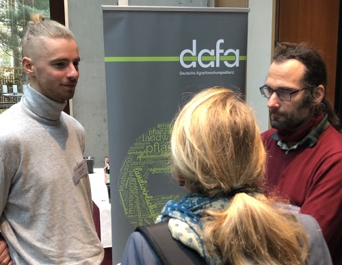 Drei Teilnehmer stehen vor einem DAFA-Plakat und reden miteinander.
